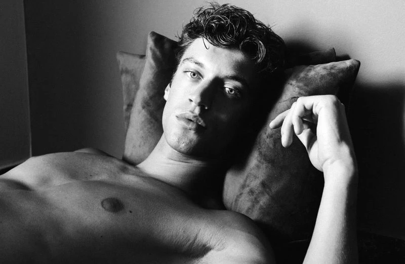 Black and white portrait of shirtless man lying down on pillow, looking at camera with serious expression in an indoor setting.