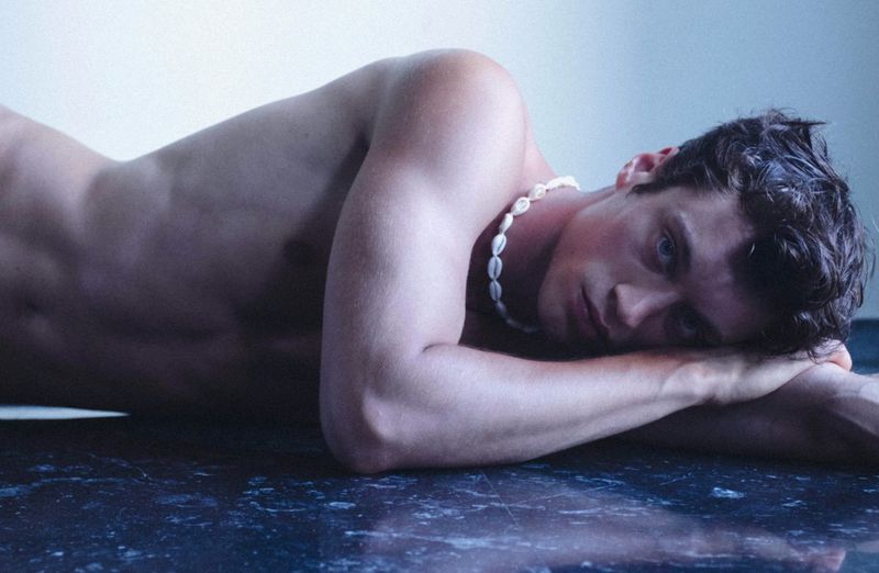 Adult male reclining on a dark surface, wearing a shell necklace, looking at the camera in a dimly lit, indoor setting.