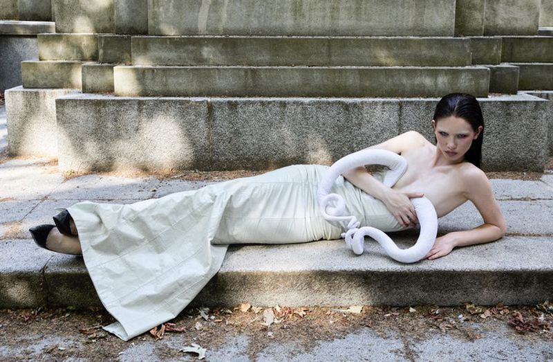 Woman in dress lies on stone steps with a white, snake-like prop in outdoor setting with soft, diffused sunlight creating a neutral colour palette.