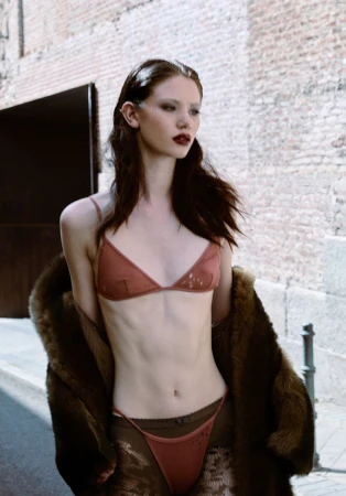 A fair-skinned adult female model poses in lingerie and a brown fur coat on an urban street with a light brick wall background.