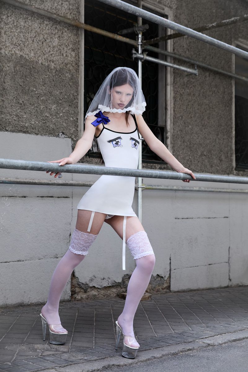 Woman poses in short white dress, lace stockings, platform heels, and a veil outside a gray building with metal scaffolding.