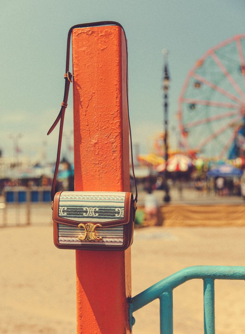 Celine crossbody bag hanging on an orange post with a blurred Ferris wheel in the background, shot outdoors on a sunny day.