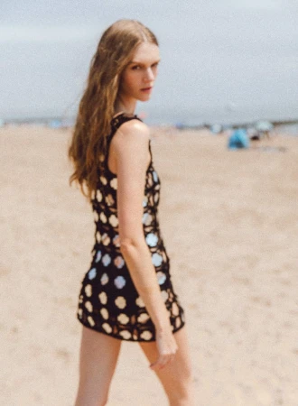Woman in a black dress poses on a sandy beach with blue sky, showcasing a blend of fashion and coastal atmosphere in an editorial shot.