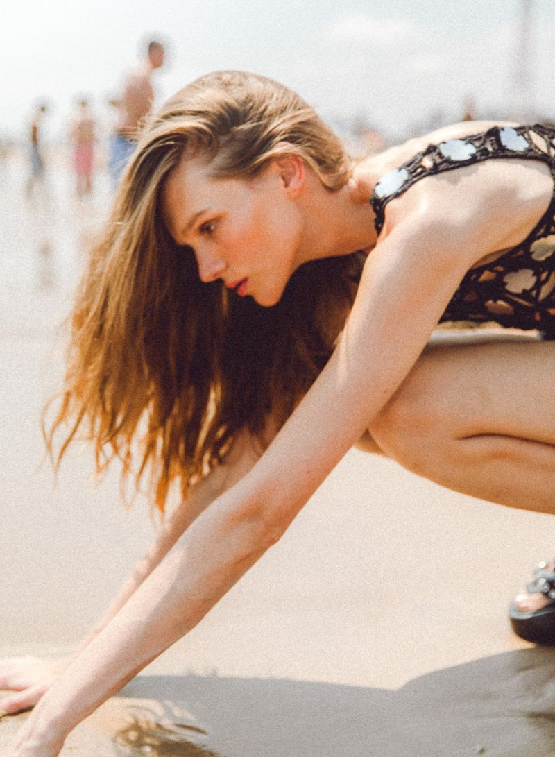 Woman in a stylish swimsuit crouches on a sandy beach on a sunny day, with other people visible in the background near the ocean.