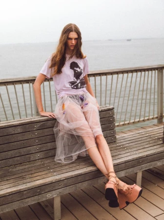 Young adult woman with long hair poses in tulle skirt and graphic tee on wooden bench at the oceanfront on a cloudy day.
