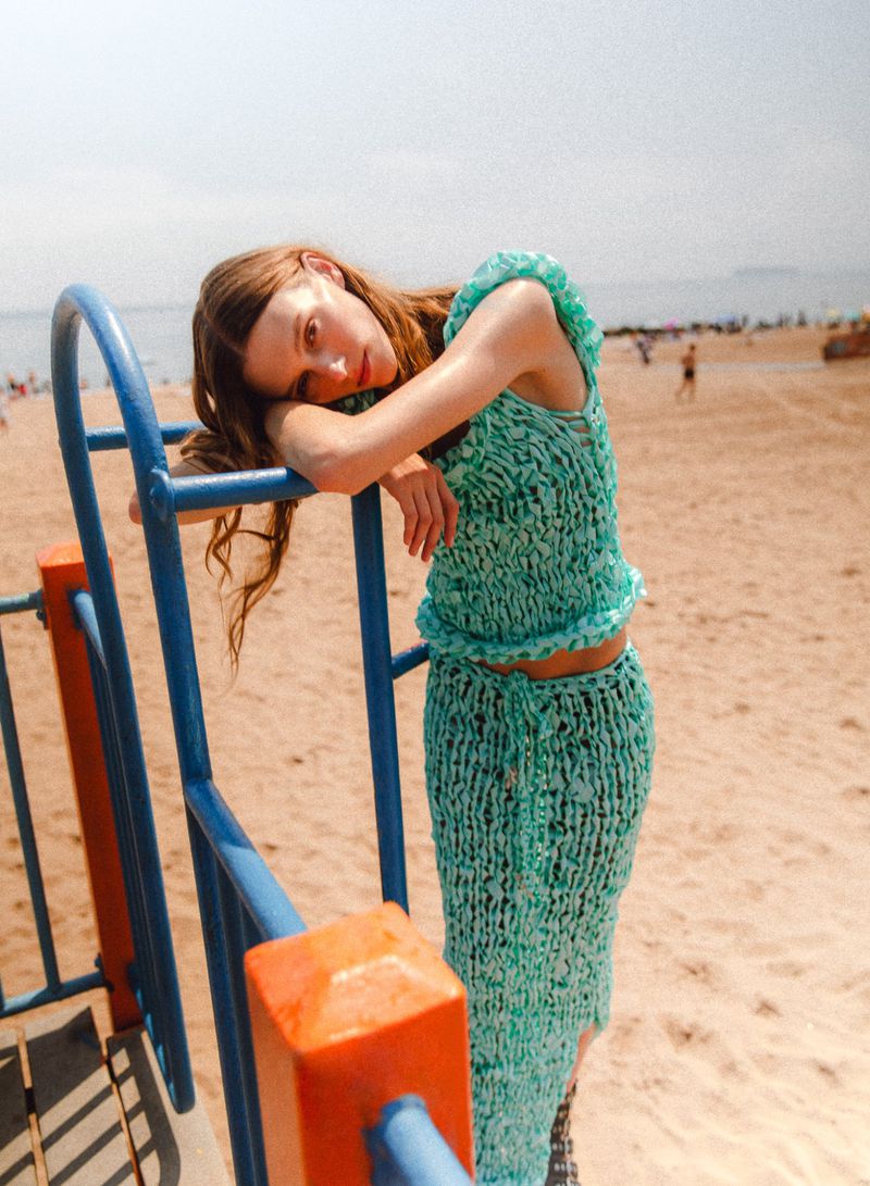 A slender woman in a knit outfit leans on a blue metal structure at the beach with other people in the distance during the daytime.
