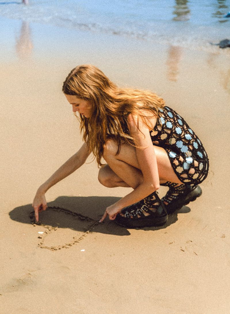 Young woman in black dress squats on a sandy beach drawing a heart shape in the sand with her fingers. Ocean waves in background.