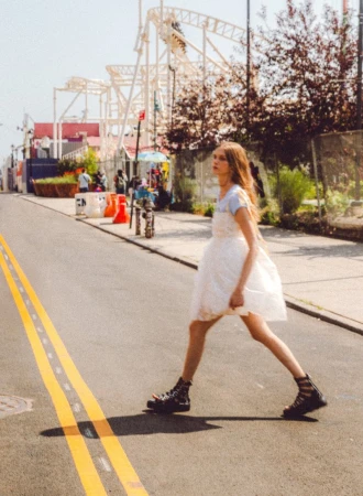 A young adult woman in a white dress walks across a street with yellow lines in an outdoor urban setting on a sunny day.