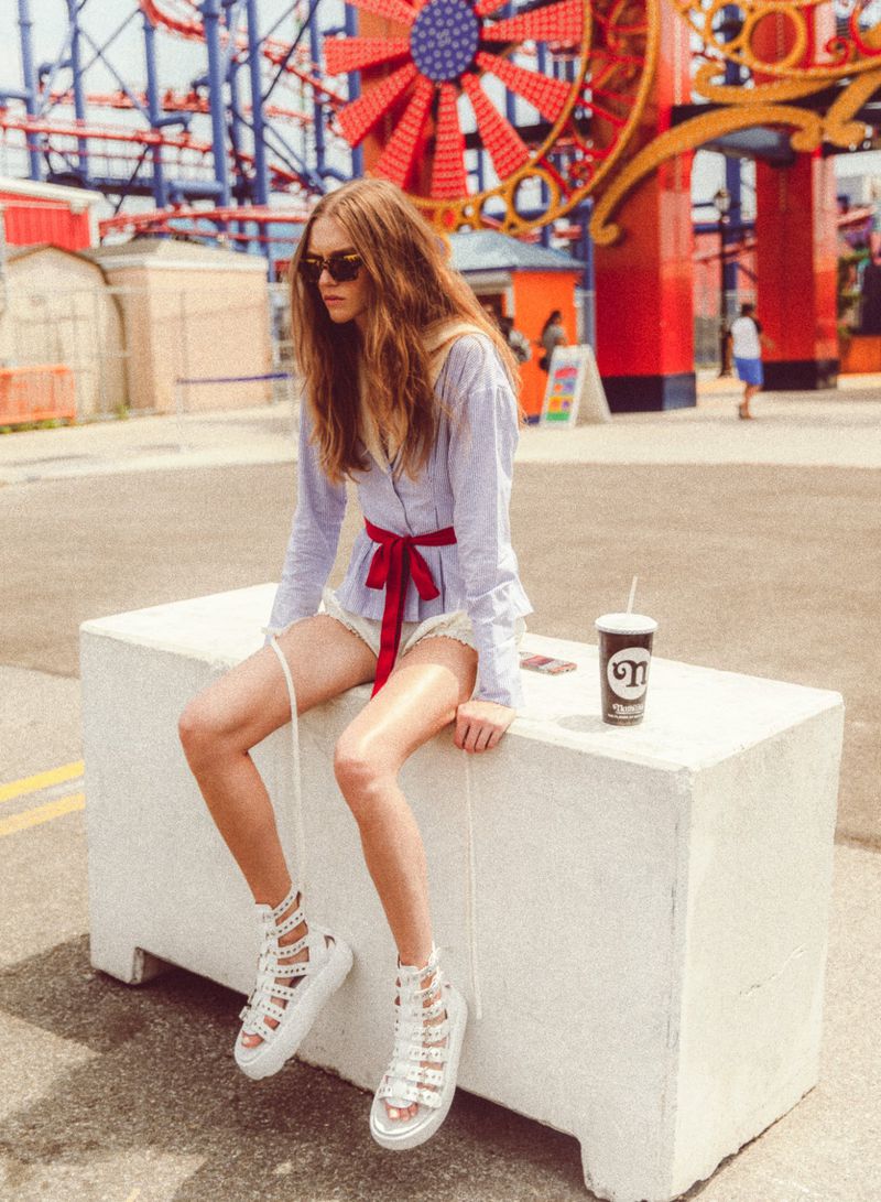 Young woman in stylish summer clothes sits on concrete block with drink in amusement park with Coney Island Cyclone rollercoaster.