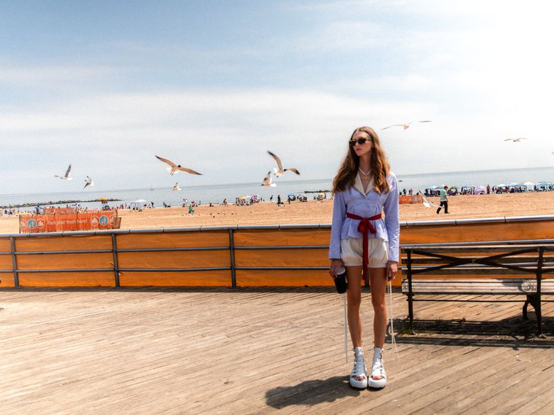 Young woman in white shorts and a blue jacket poses on a boardwalk near a beach with seagulls and people on a sunny day.