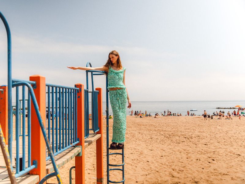 A woman in a patterned outfit stands on a playground ladder with a crowded beach and ocean in the background on a sunny day.