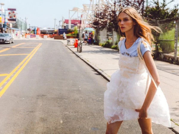Fashion photo of a woman in a white dress crossing a city street near a boardwalk with a fairground during the daytime.