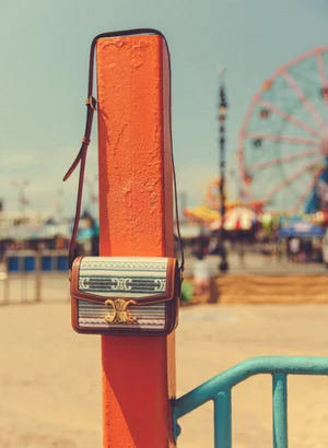 A patterned leather and canvas handbag hanging from an orange post with an amusement park and ferris wheel in the blurred background.