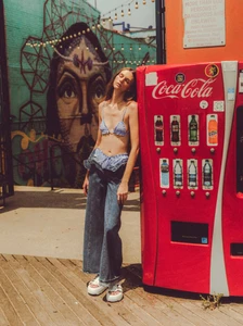 Model wearing a blue patterned bikini top and wide-leg denim jeans leaning against a bright red vending machine.