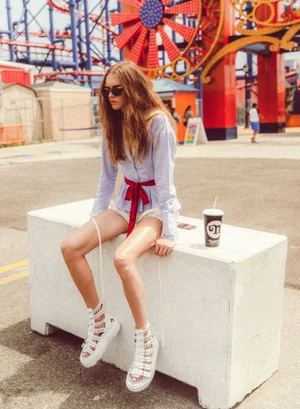 Model wearing blue striped blouse and white platform sandals sitting on a white block with amusement park background