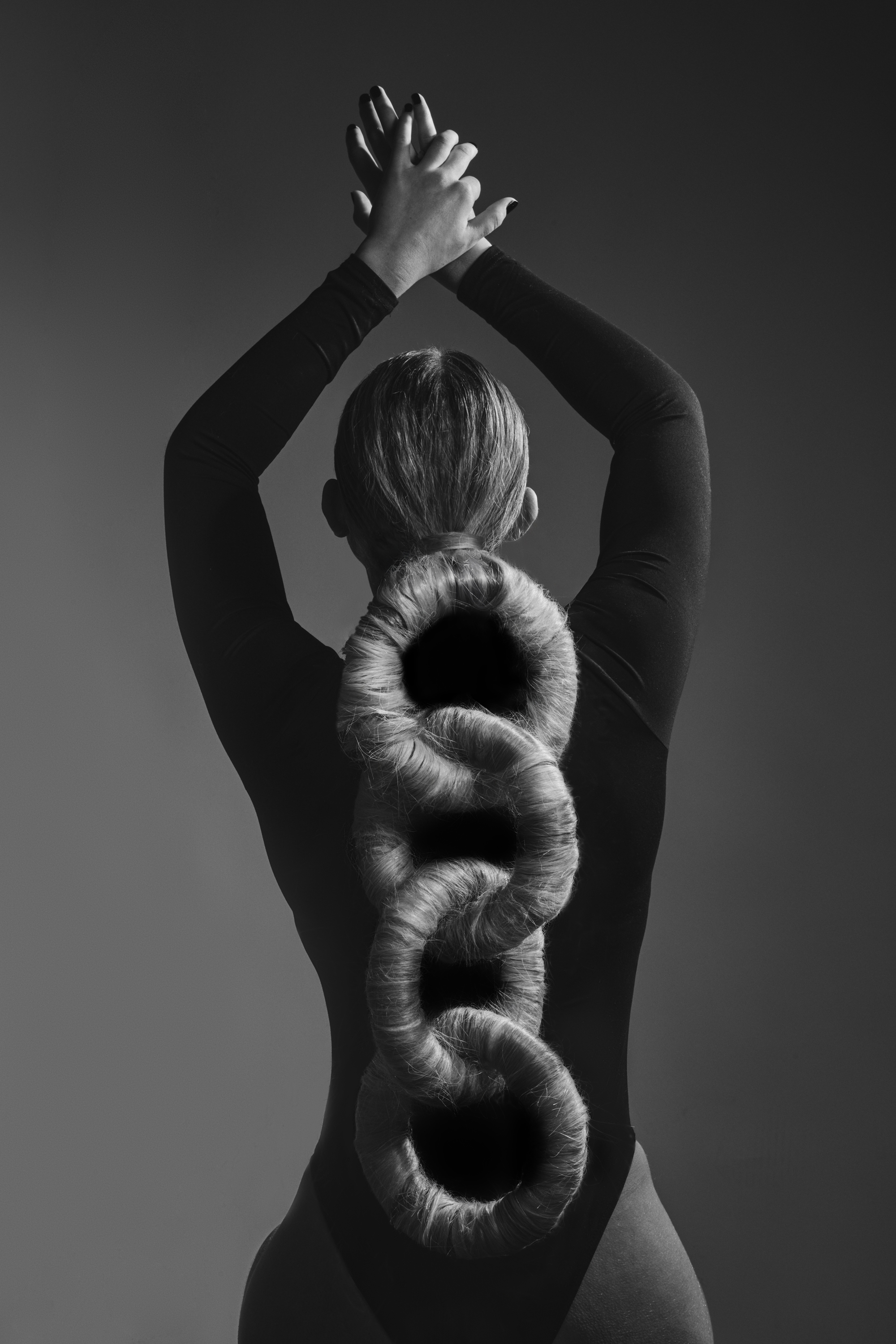 Back view of a female with a blonde hair braid formed into interconnected rings, wearing a black leotard against a grey background.