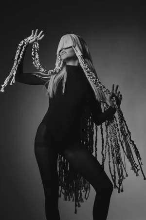 Model with long blonde hair and braided hair extensions wearing a black bodysuit against a grey studio background