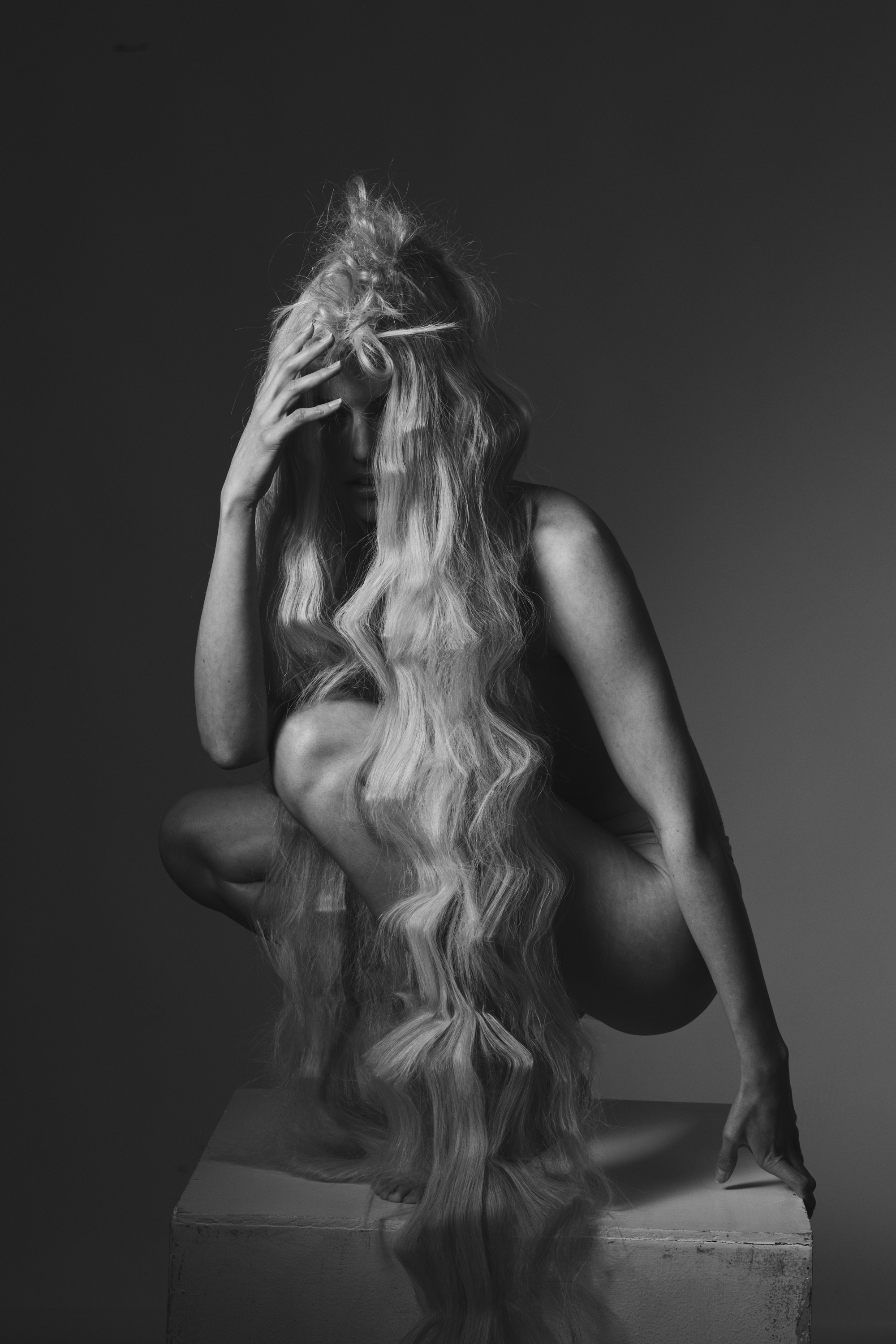 Monochrome portrait of a crouched woman with long blonde hair obscuring her face, seated on a white box against a grey background.