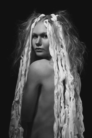 Monochrome portrait of a young adult woman with an elaborate headpiece made of fabric strips, looking over her shoulder in a studio setting.