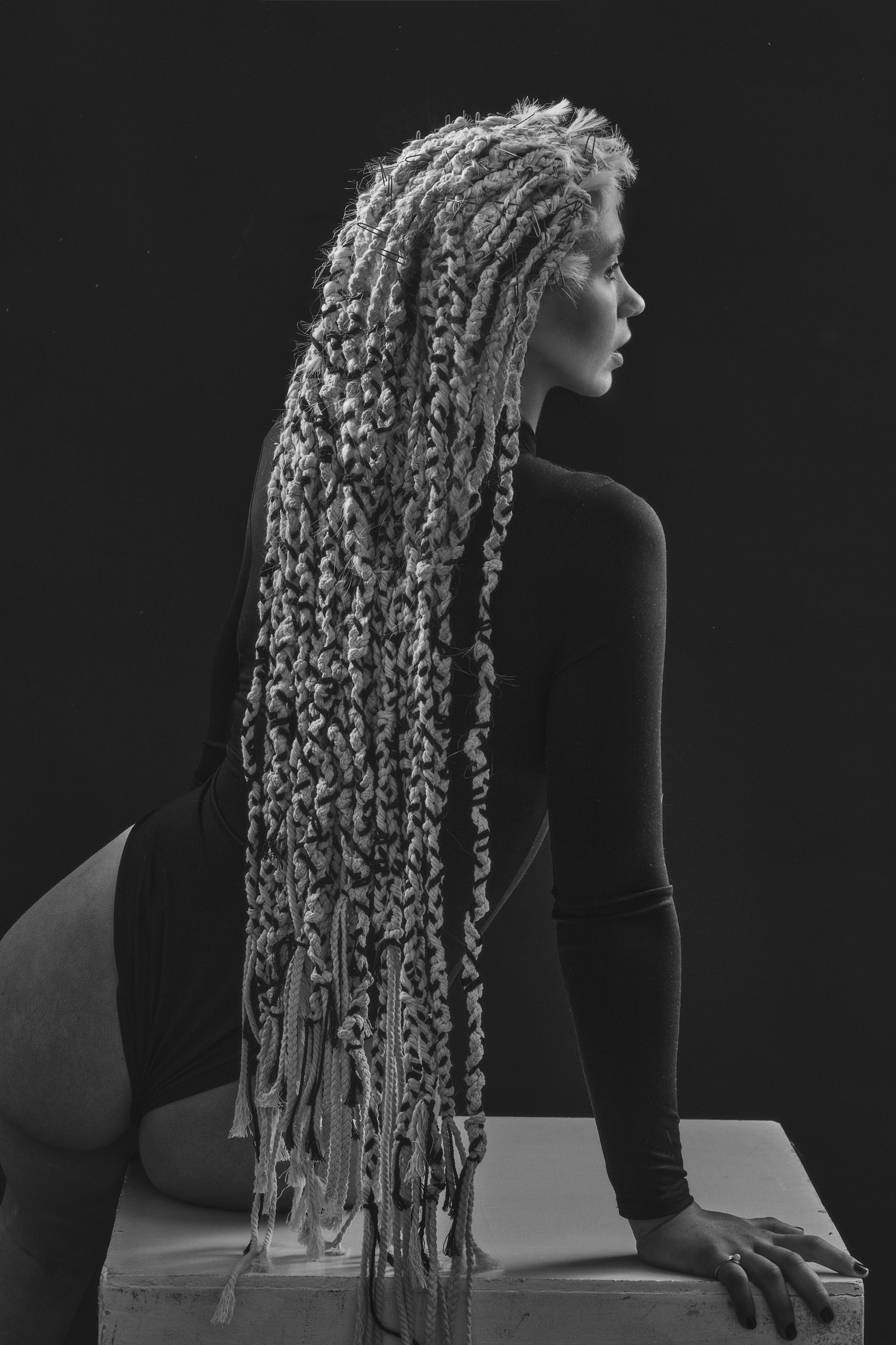 Monochrome portrait of a woman with long textured dreadlocks wearing a dark bodysuit, leaning on a white block against a black backdrop