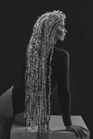 Monochrome portrait of a woman with long textured dreadlocks wearing a dark bodysuit, leaning on a white block against a black backdrop