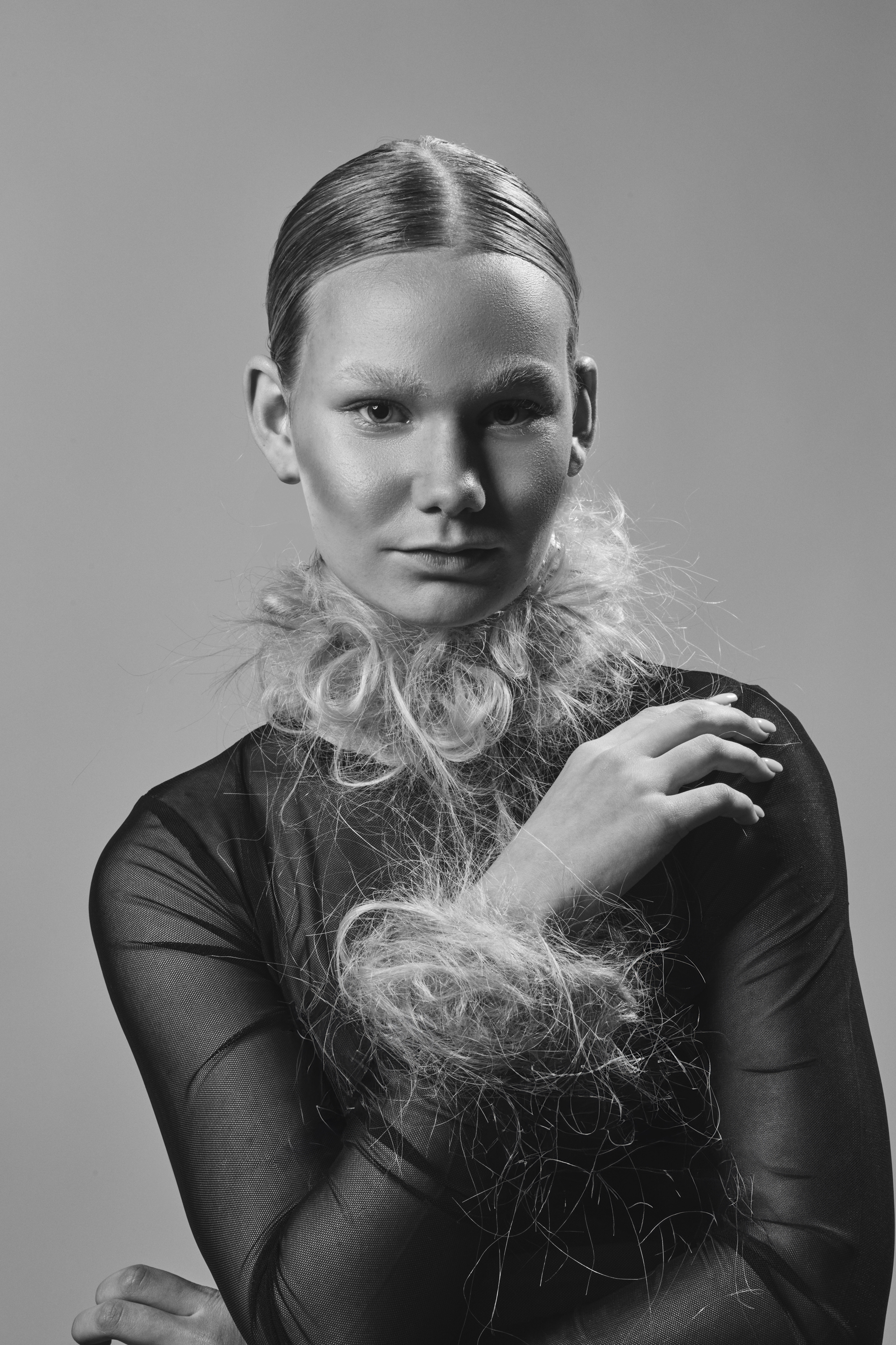 Black and white portrait of an adult woman with her hand near her face, wearing a sheer top with blonde fluffy collar and cuffs.