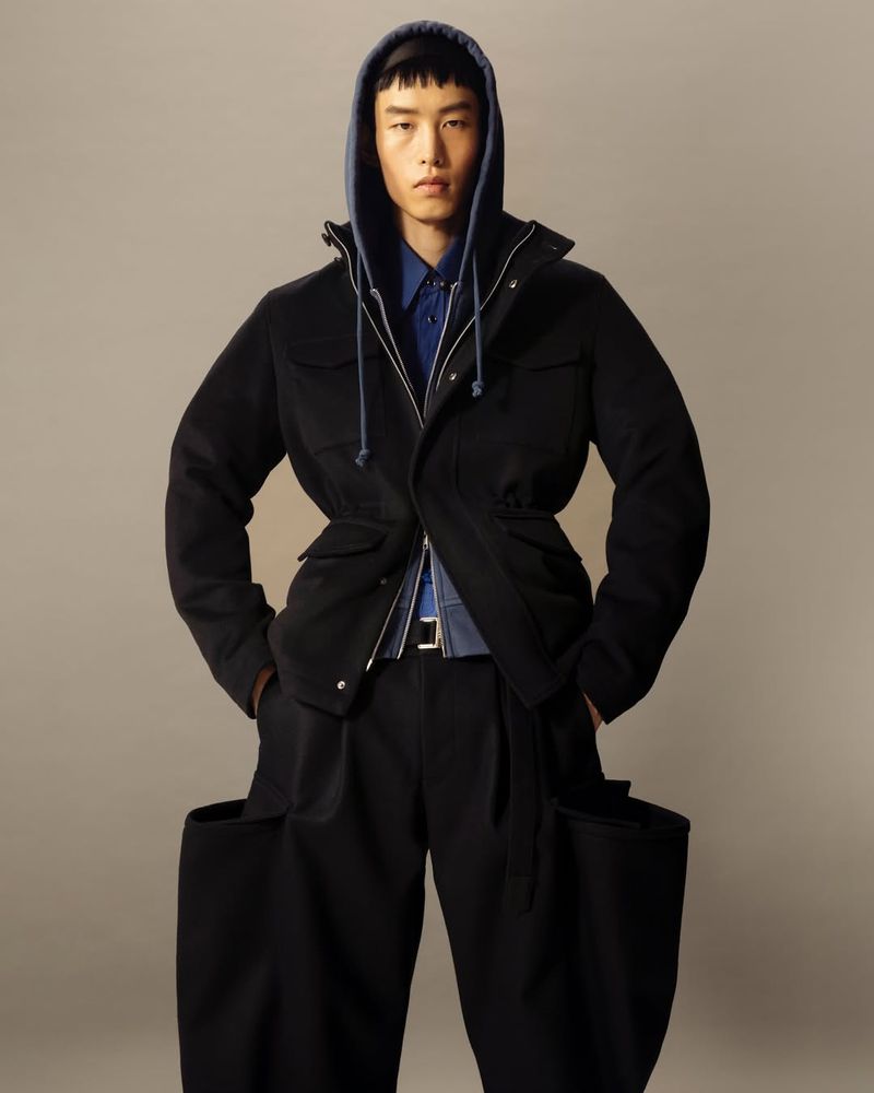 Asian male model wearing a blue hooded shirt, black jacket, belt, and wide-legged pants posing in a studio with a neutral background.