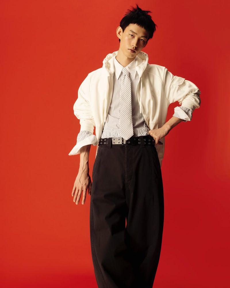 Stylish Asian man in a white jacket, striped shirt, and wide-leg pants poses against a vibrant orange background in a studio fashion shot.