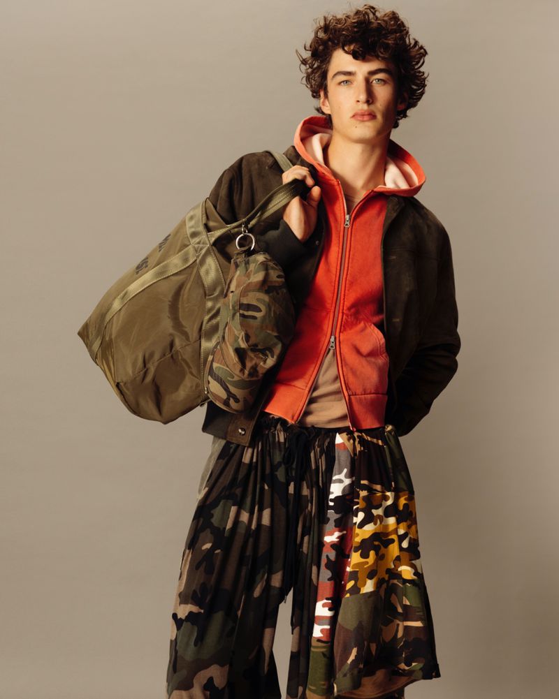 Studio shot of a young male fashion model wearing a hooded jacket, camouflage pants, and carrying a duffel bag in muted tones.