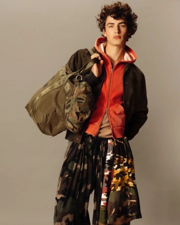 Studio shot of a young male fashion model wearing a hooded jacket, camouflage pants, and carrying a duffel bag in muted tones.