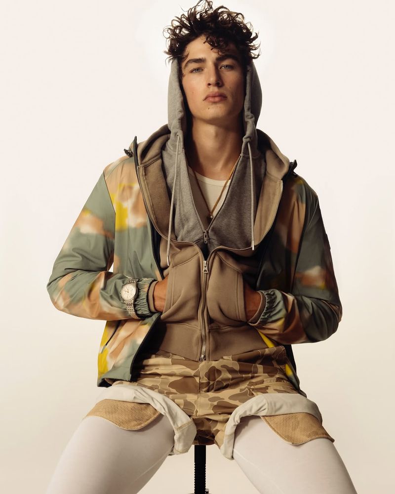 Male fashion model in a hooded jacket and camo shorts sits in a studio with a neutral backdrop for a modern editorial shot.