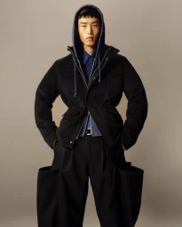Model in black jacket with hood and dark wide trousers standing against a plain neutral backdrop