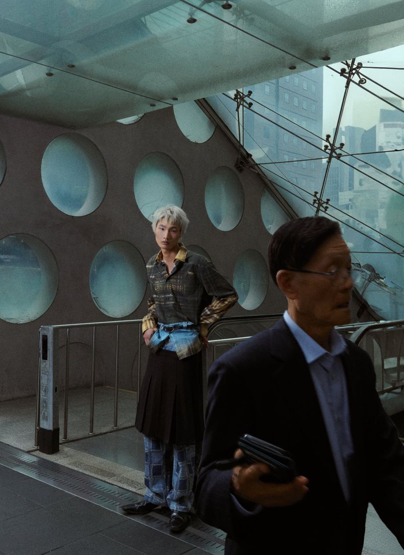A young adult in patterned shirt and kilt stands indoors near an older adult in a suit, in an urban setting with an architectural backdrop.