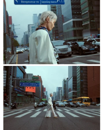 Adult with white hair crosses street in white suit in urban environment. Modern buildings and street signs in the background. 