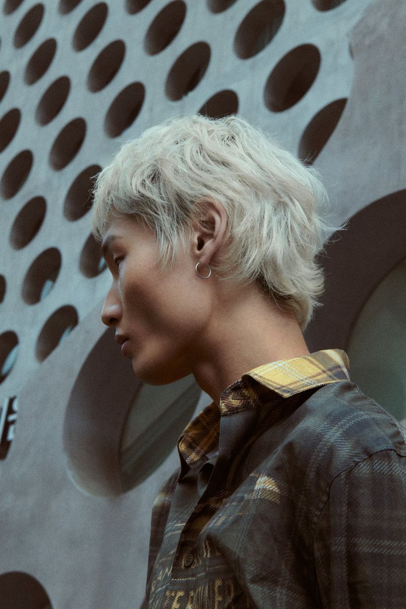 A young adult Asian man poses in a plaid shirt next to a textured wall with circular cutouts, creating a modern, urban feel.