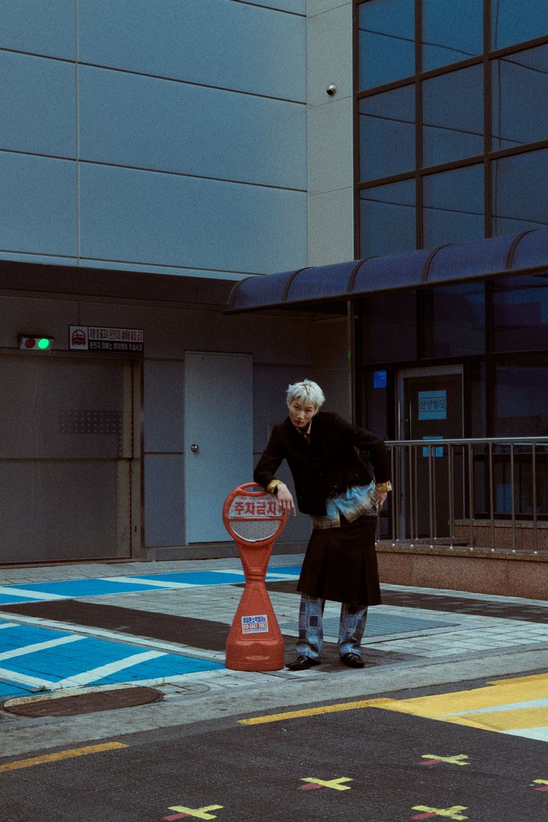A young adult stands outdoors in an urban setting next to a red parking barrier, wearing a black top and denim skirt.