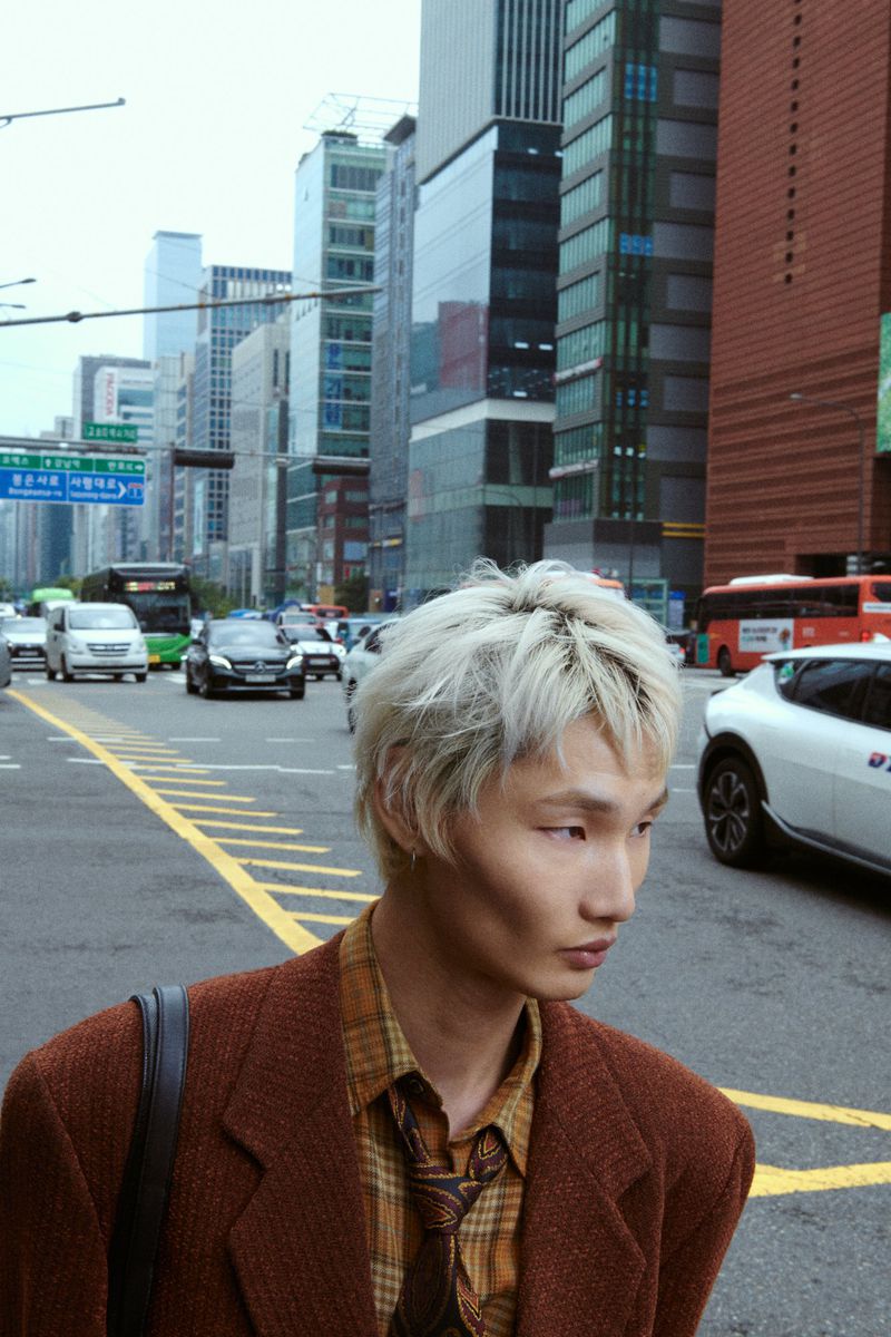 Young adult with bleached hair in a brown blazer standing on a city street with tall buildings and vehicle traffic in the background.