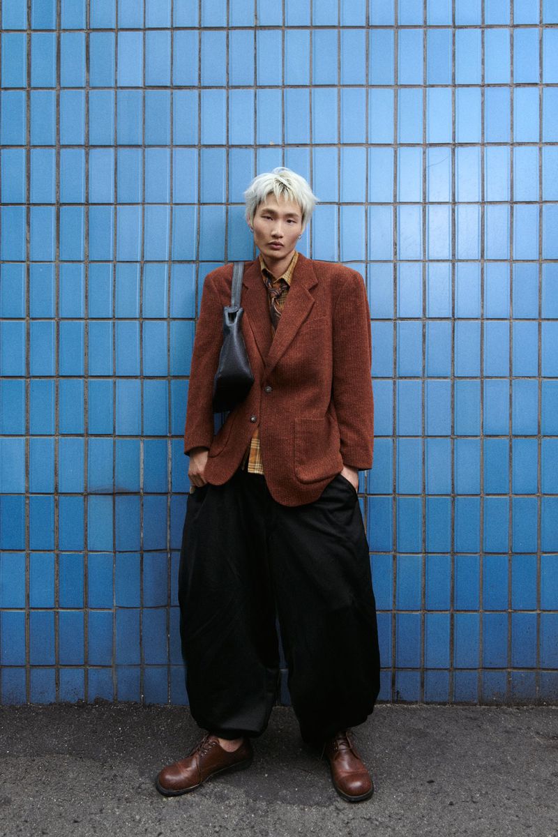 Young man with light hair wearing a brown tweed blazer, black pants and leather shoes standing in front of a blue tiled wall.
