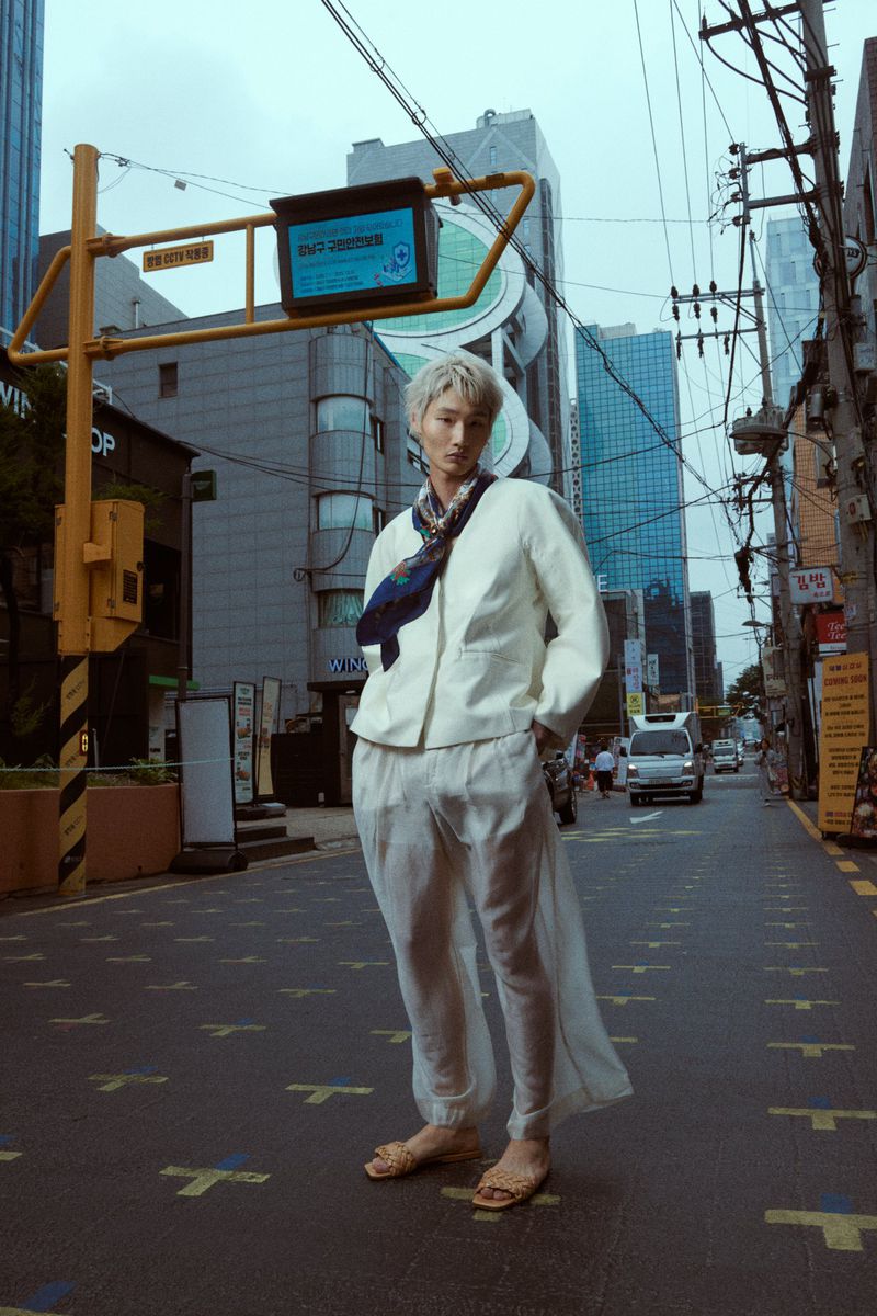 Full-length shot of an adult male with blonde hair in a white jacket, sheer pants and sandals, standing on a patterned city street.