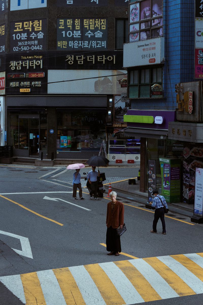 Adult female with short gray hair stands on a crosswalk, holding a briefcase, city street background, umbrellas and signage.
