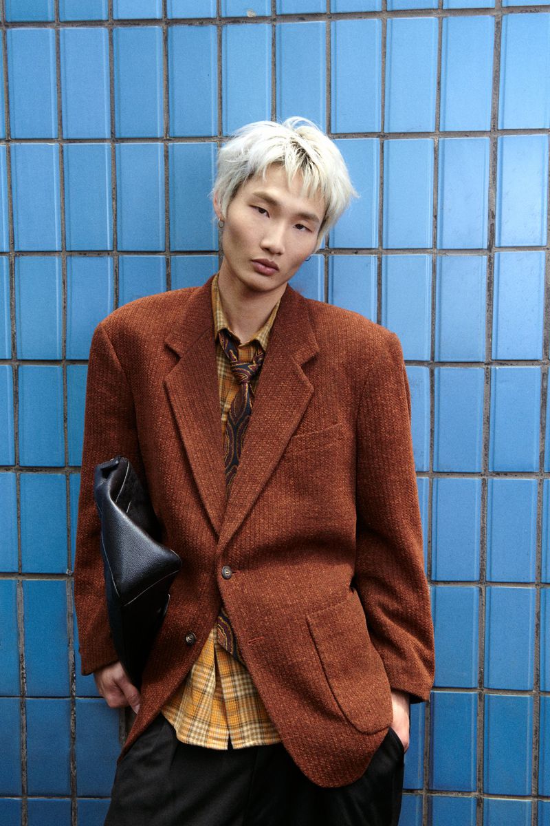 Adult male model with light hair poses against a blue tiled wall, wearing a brown blazer, yellow shirt, black pants, and a black leather bag.