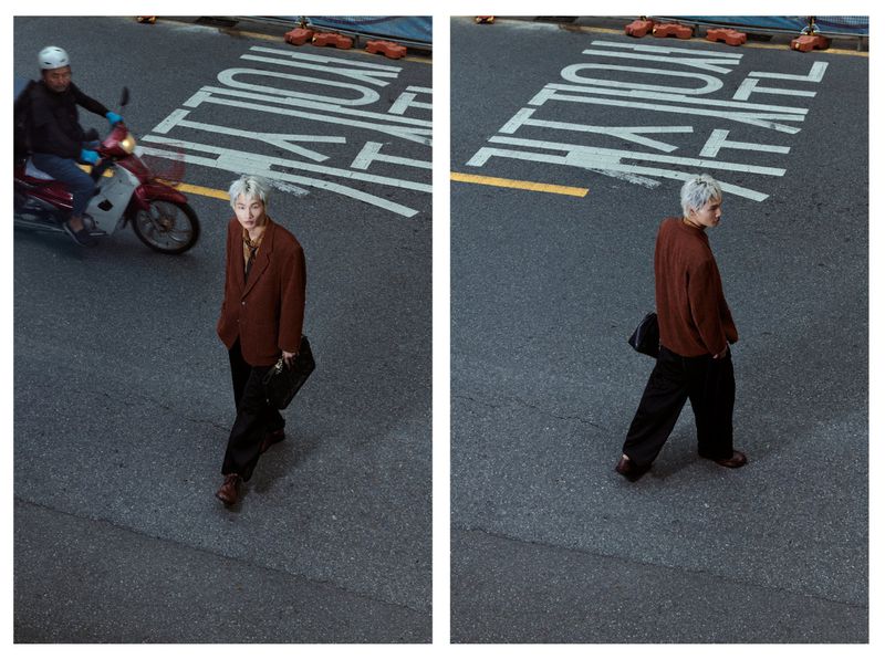 Stylish man in brown blazer walks across asphalt street with painted crosswalk markings and a motorbike passing in the background.