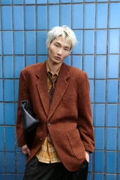 Model with platinum blonde hair wearing a rust brown blazer and patterned shirt holding a dark clutch against blue tiled wall