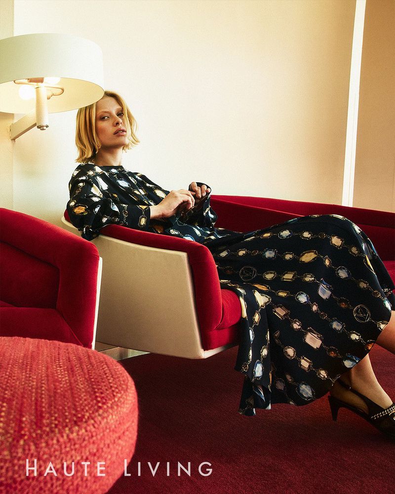 Woman sits in a red chair wearing a long-sleeve dress with a chain print.  A mid-century style lamp illuminates the interior space.