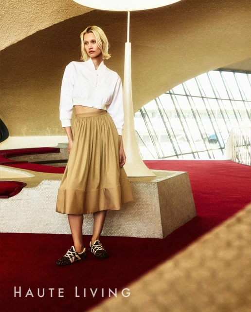 A fair-skinned female adult model in a white blouse and khaki skirt poses inside a modern building with red flooring and unique architecture.