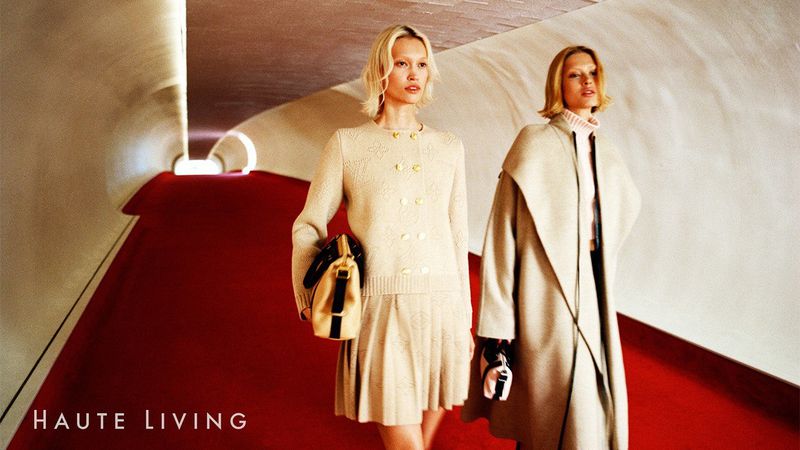 Two blonde fashion models pose in an architectural space, wearing light neutral knitwear and coats, with a red floor and neutral walls.