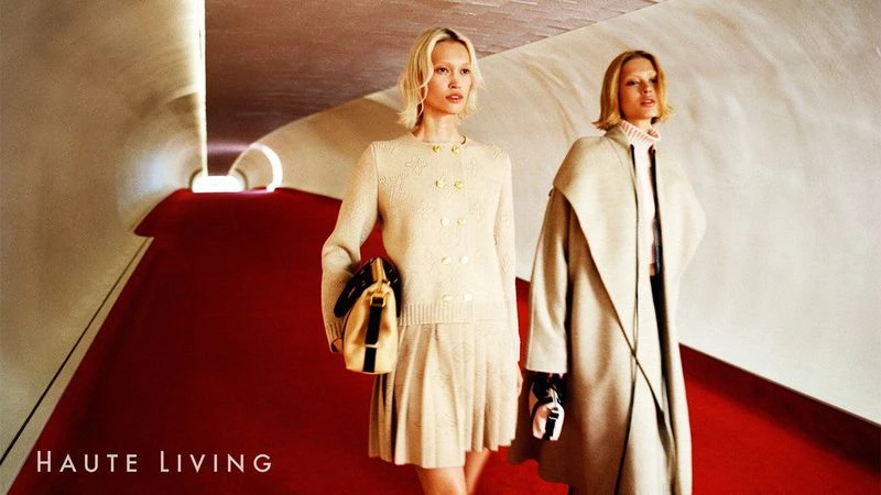 Two blonde fashion models pose in an architectural space, wearing light neutral knitwear and coats, with a red floor and neutral walls.