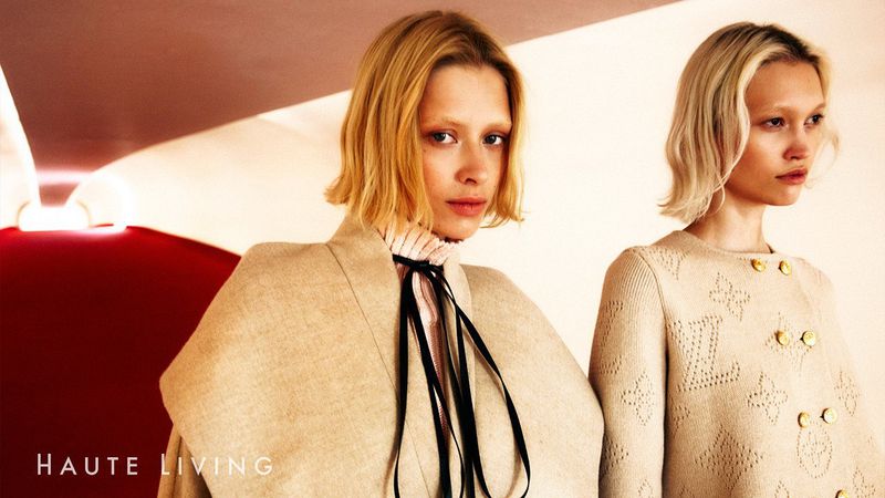 Two blonde fashion models pose indoors wearing beige patterned sweaters with red and beige background. Haute Living text is visible.