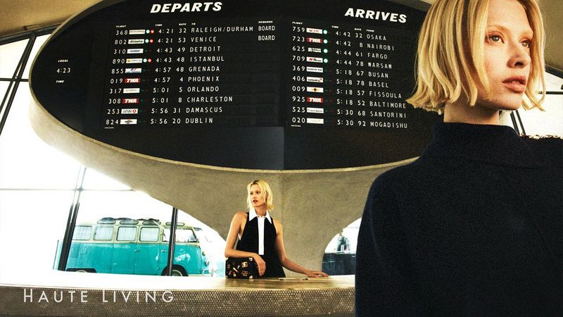 Two women posing indoors near an airport-style departure board, with a vintage van visible in the background and a dark navy sweater nearby.
