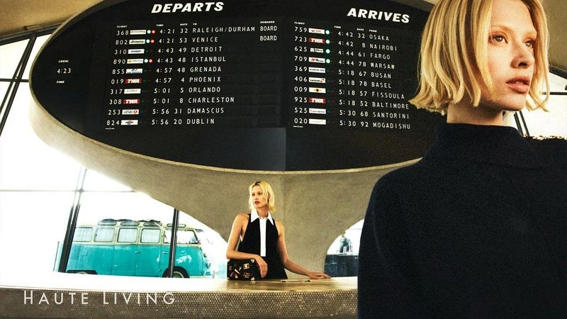 Two women posing indoors near an airport-style departure board, with a vintage van visible in the background and a dark navy sweater nearby.